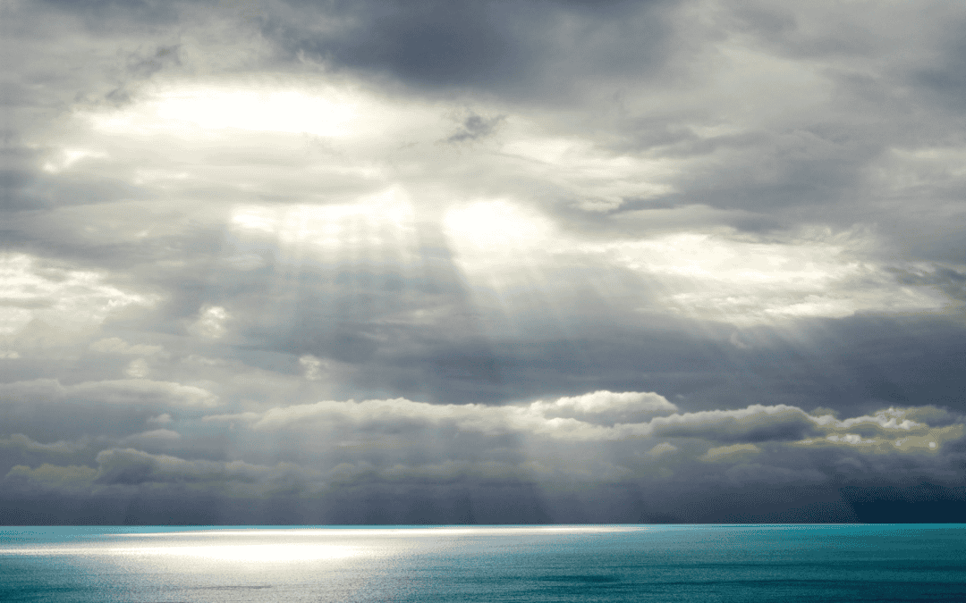 Es sind viele Wolken und ein ruhiges Gewässer zu sehen. Zwischen den dunklen Wolken kommen helle Strahlen hervor und scheinen auf das Wasser